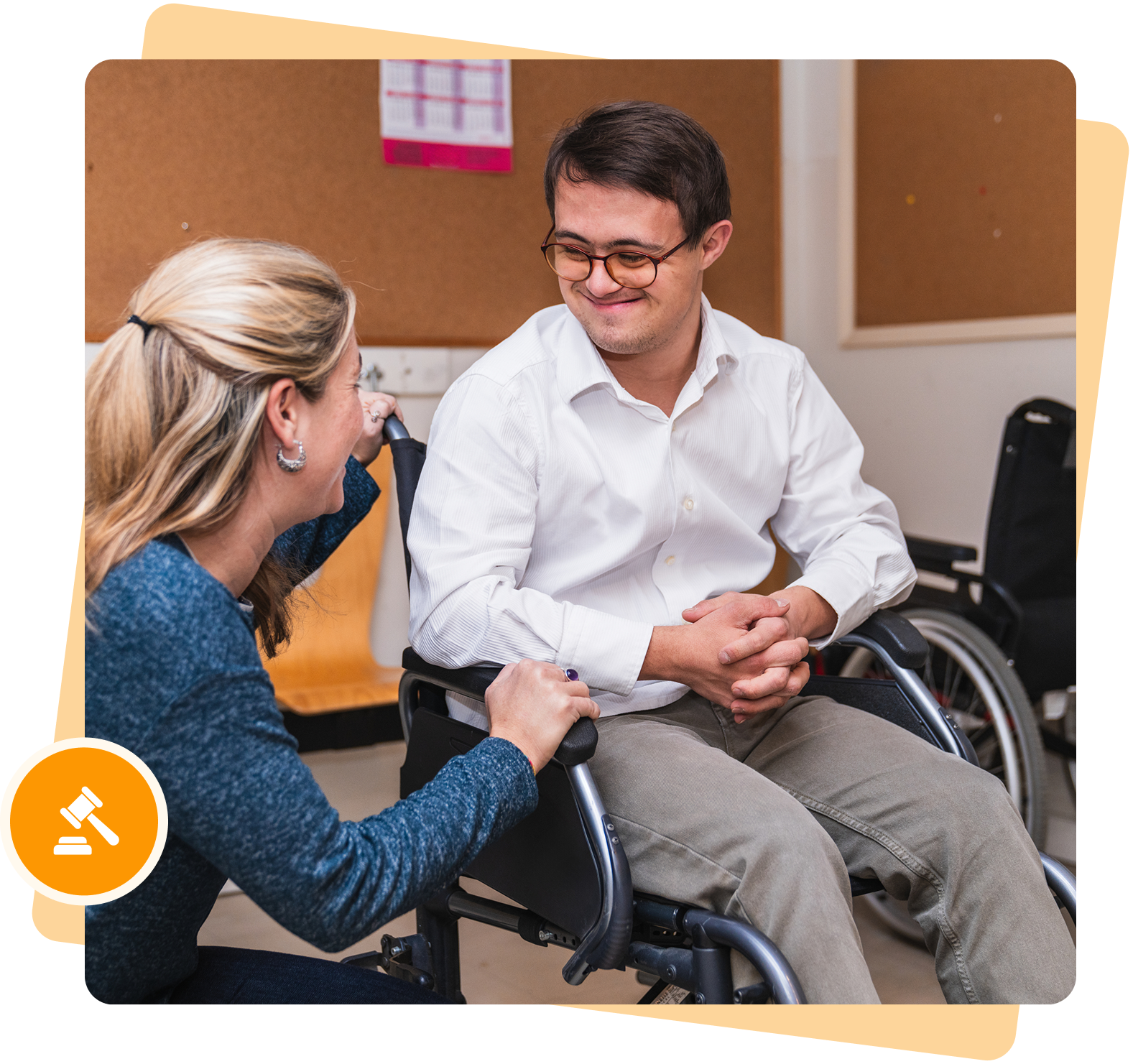 Group 1321316692 Two people are talking, one seated in a wheelchair and the other crouching beside. They are in a room with bulletin boards.