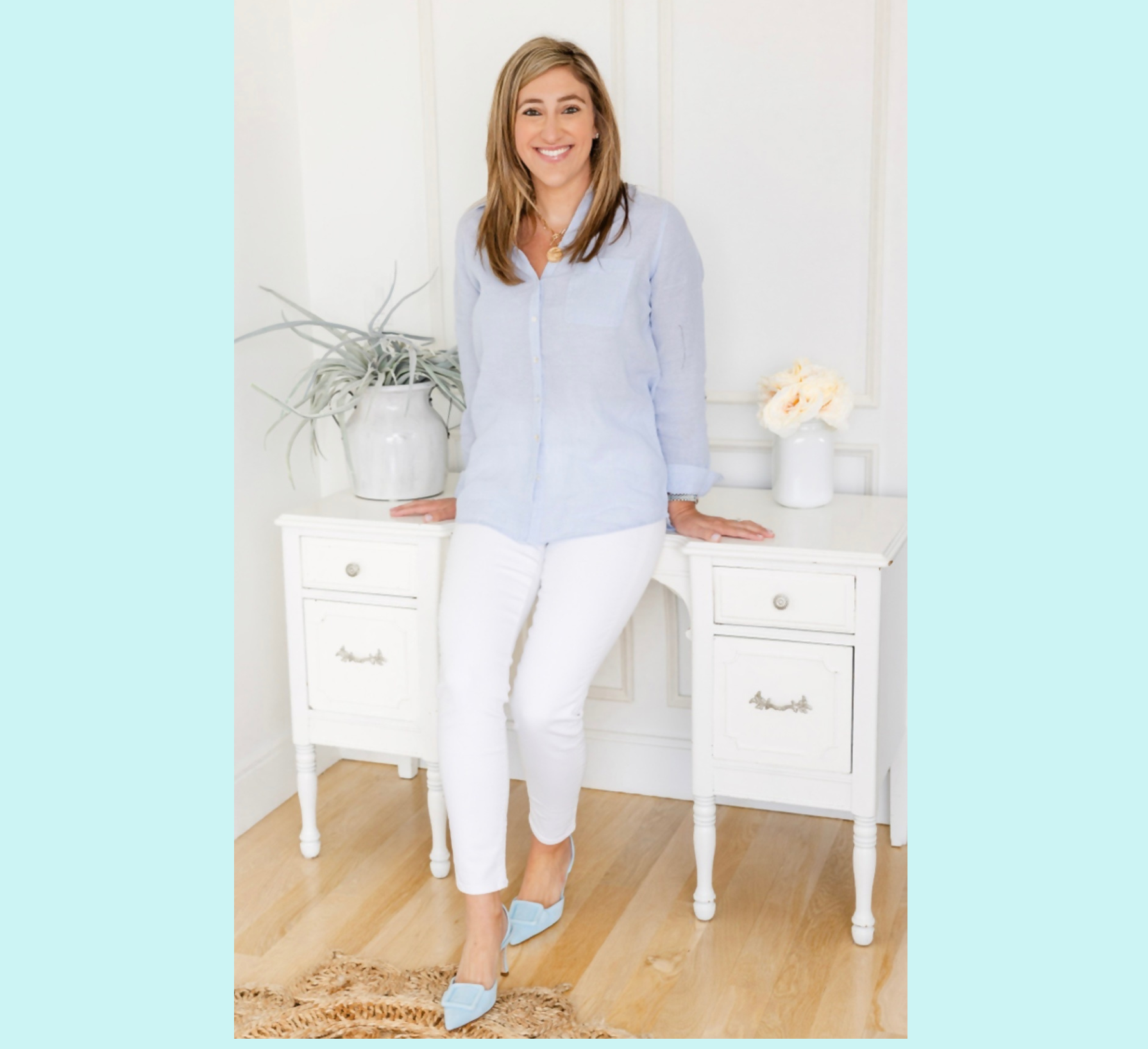 Untitled design (1) A woman in a light blue shirt and white pants stands, smiling, in front of a white table with a plant and a vase of flowers in a bright room with light wood floors.