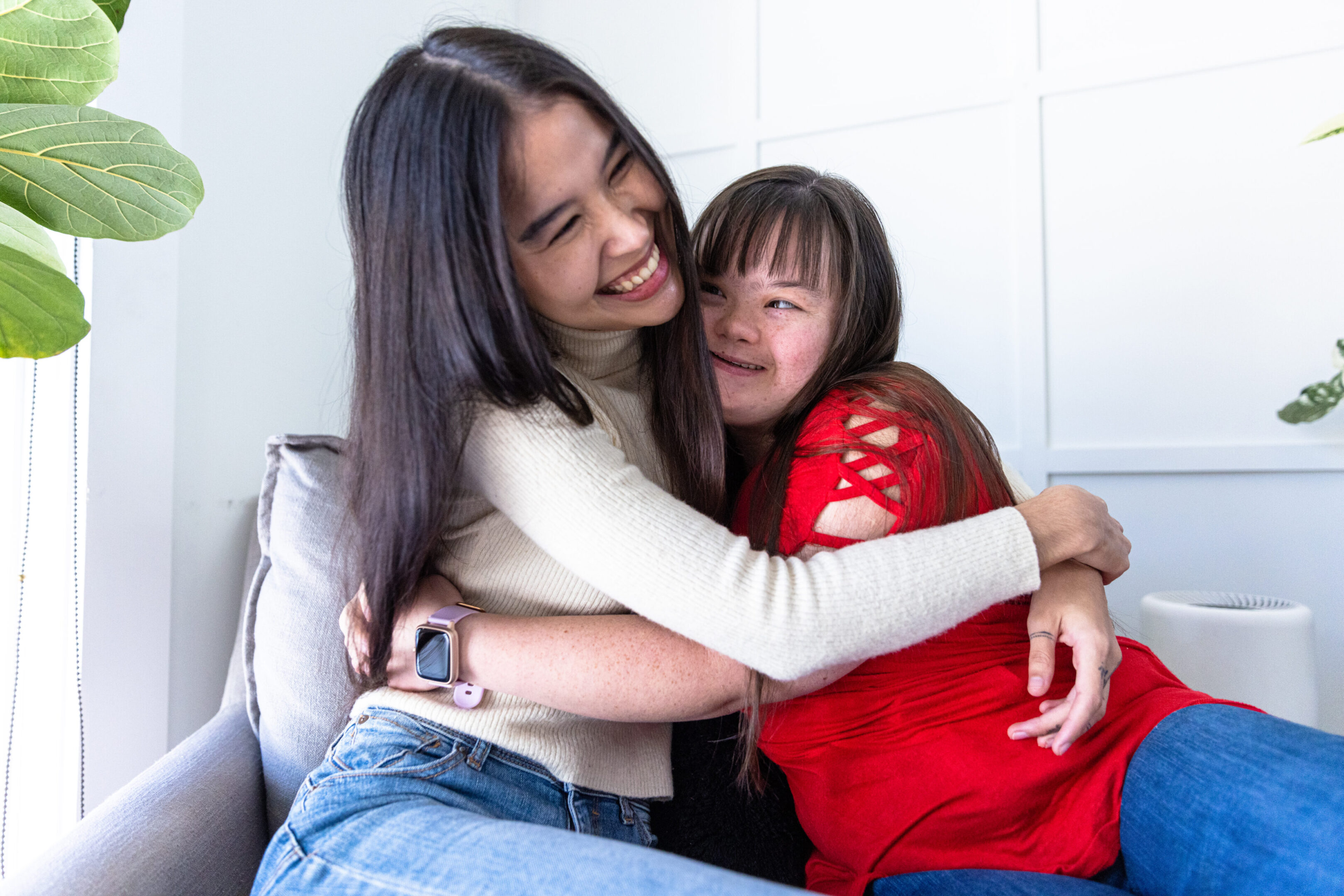 Sisters lovingly hugging on the comfy chair at home. One sister has Down's syndrome. Two women seated on a couch share a warm hug and smile. One has long dark hair, the other is wearing a red top. Bright room with green plant on the side.